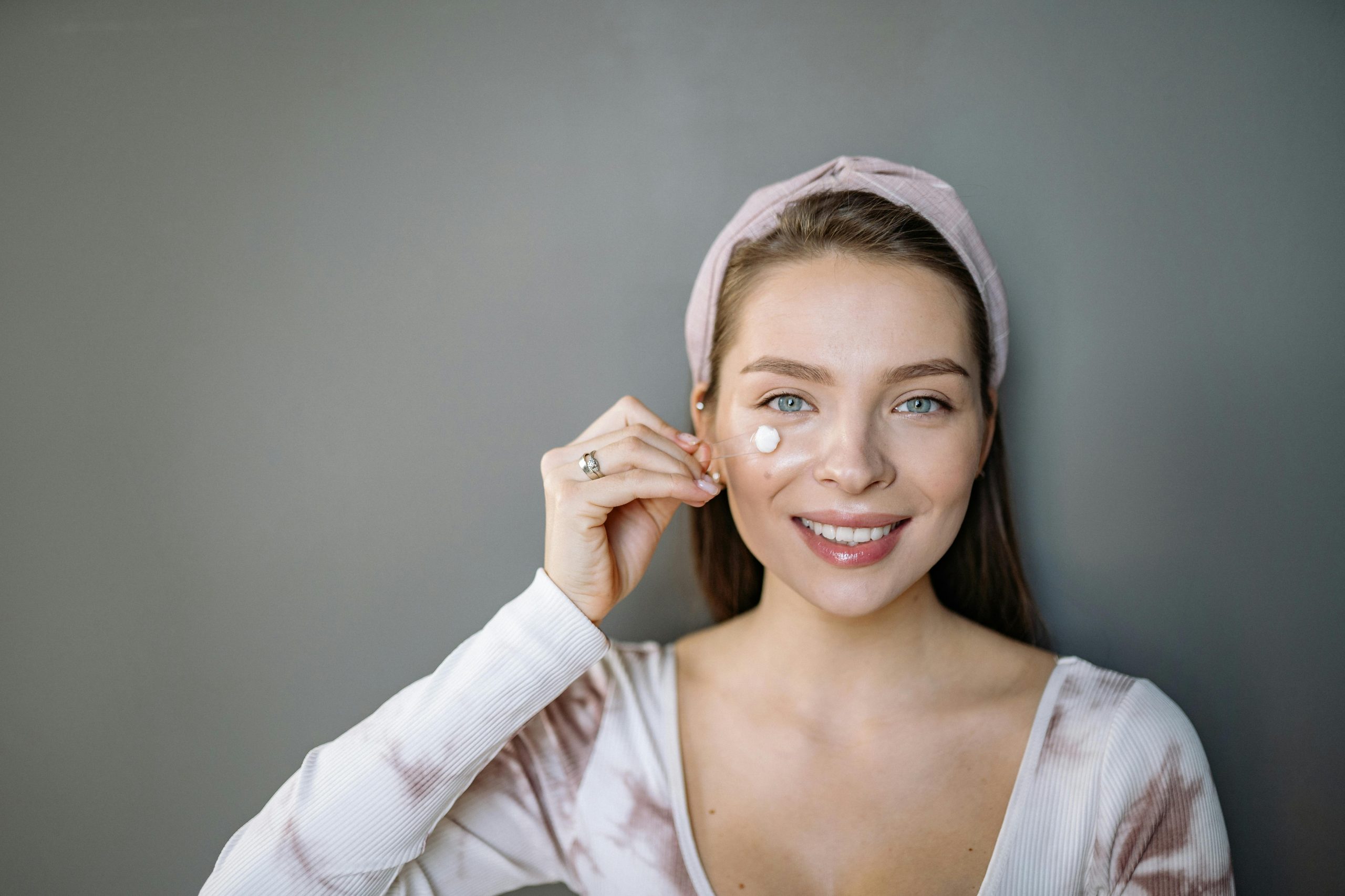 A woman with gray eyes and headband applying skincare cream indoors, smiling. Une femme aux yeux gris et bandeau appliquant une crème de soins de la peau à l'intérieur, souriante.