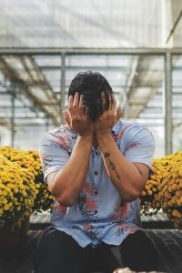 A man covering his face, sitting among potted yellow flowers in an urban greenhouse. Un homme se couvrant le visage, assis parmi des fleurs jaunes en pots dans une serre urbaine.