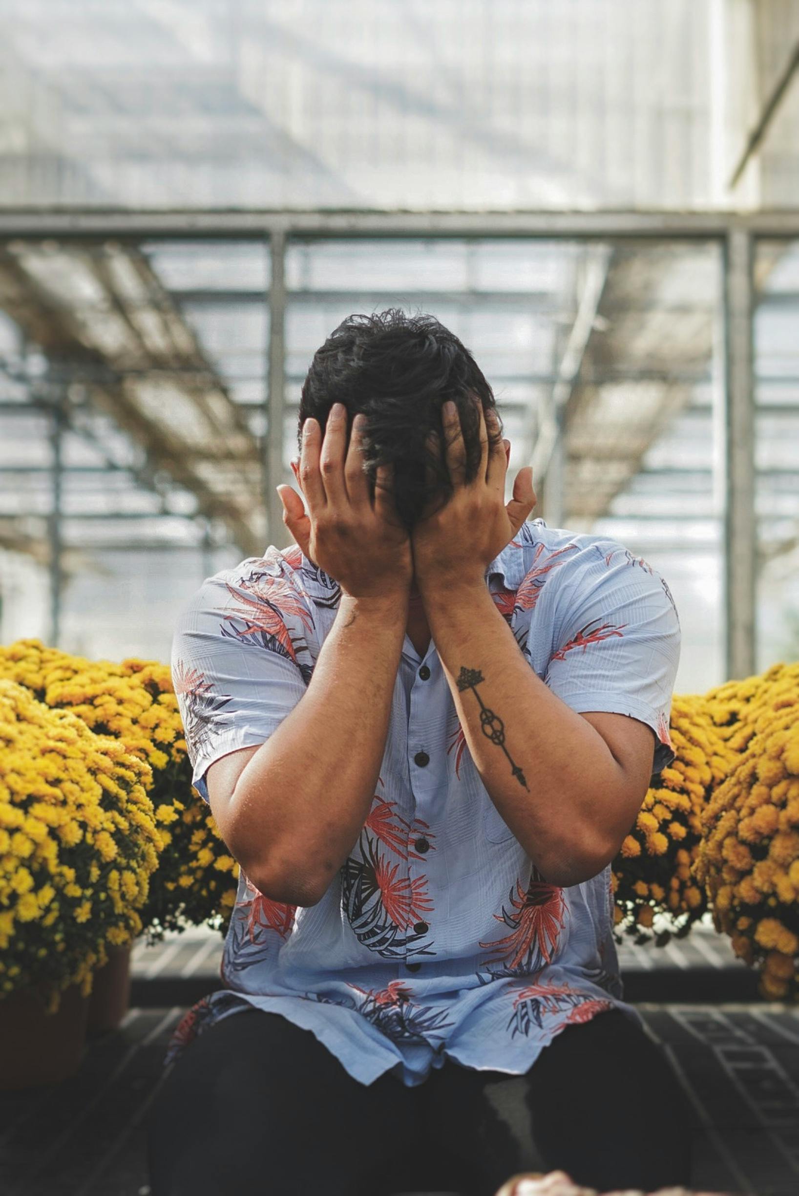 A man covering his face, sitting among potted yellow flowers in an urban greenhouse. Un homme se couvrant le visage, assis parmi des fleurs jaunes en pots dans une serre urbaine.