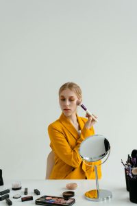 A woman in a yellow blazer applying makeup with a brush in front of a mirror indoors. Une femme en blazer jaune appliquant du maquillage avec un pinceau devant un miroir en intérieur.