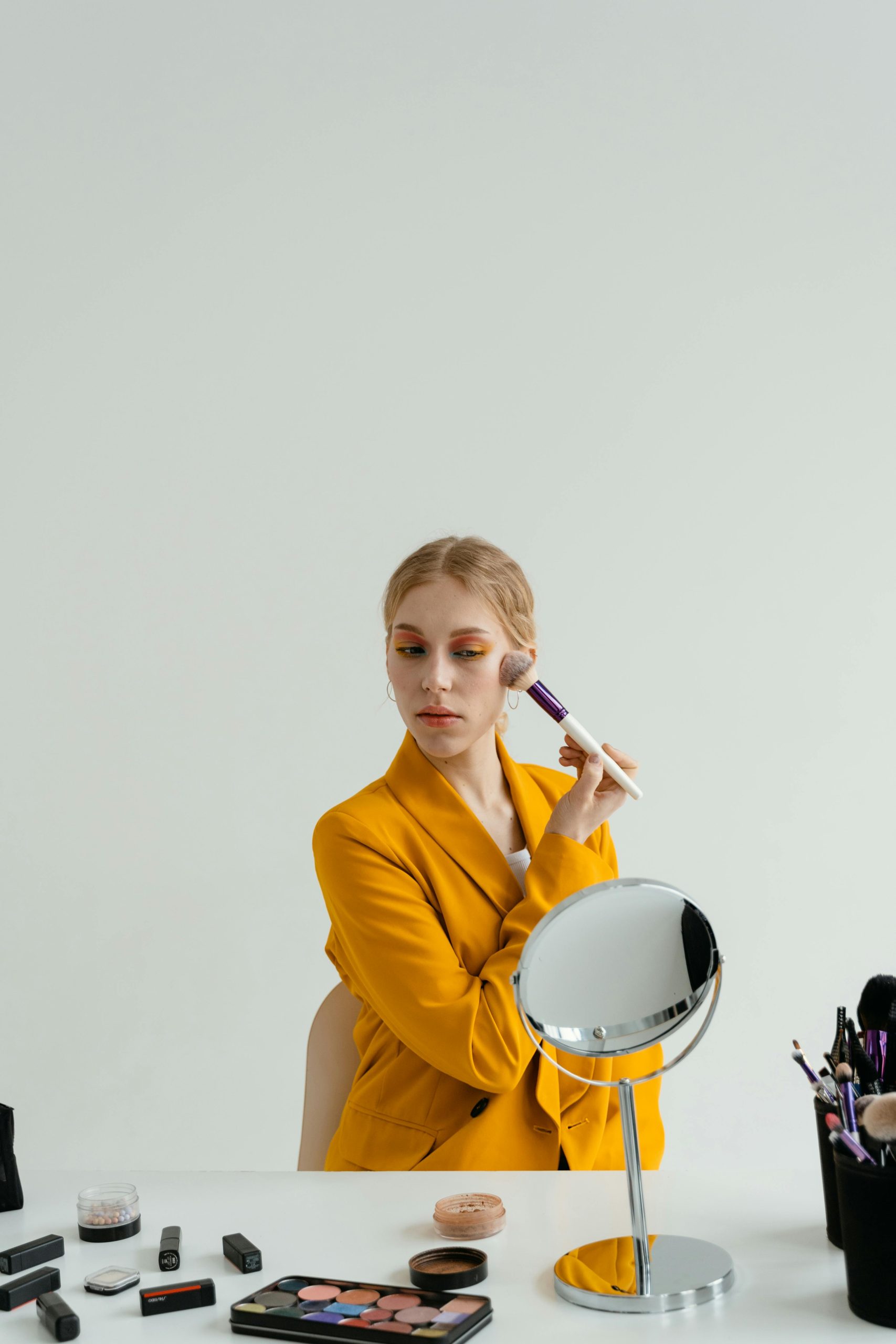 A woman in a yellow blazer applying makeup with a brush in front of a mirror indoors. Une femme en blazer jaune appliquant du maquillage avec un pinceau devant un miroir en intérieur.