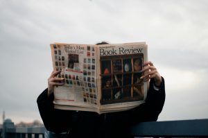 Individual holding a book review newspaper with colorful nails against a cloudy sky backdrop. Une personne tenant un journal d'examen de livre avec des ongles colorés sur fond de ciel nuageux.