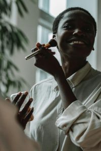 Femme noire souriante en chemise boutonnée appliquant du maquillage au pinceau à l'intérieur.