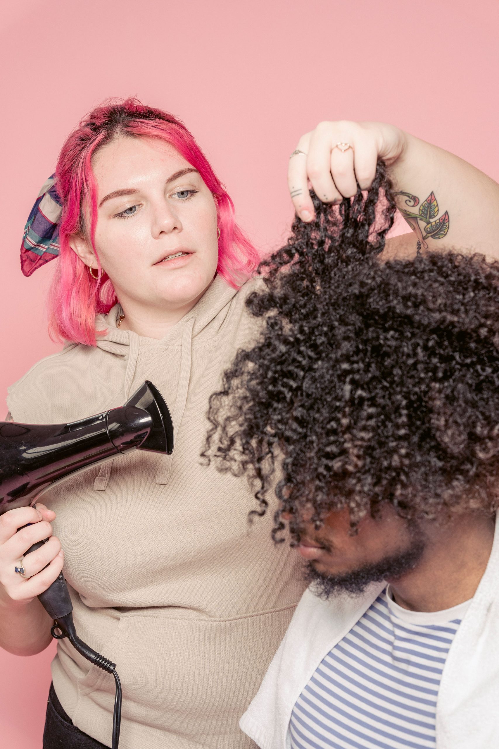Coiffeuse replète concentrée séchant les cheveux Afro noirs d'un client homme afro-américain sur fond rose dans un studio lumineux moderne.