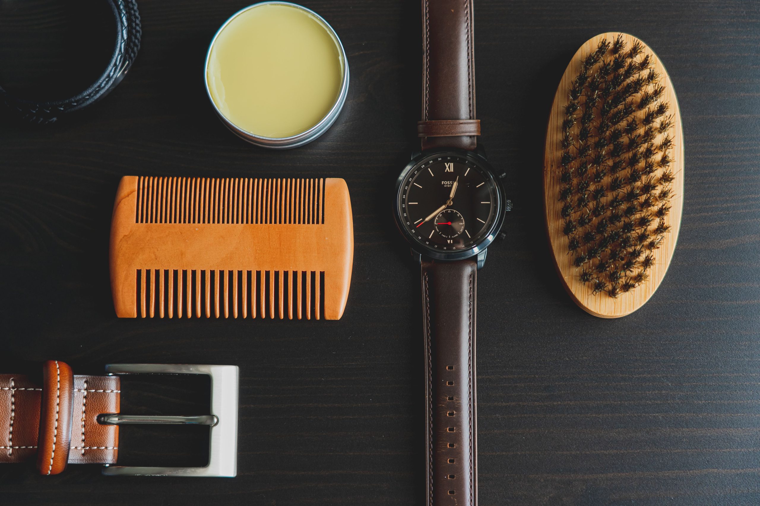 Flat lay of men's grooming accessories including watch, brush, comb, and pomade. Flat lay de accessoires de toilettage pour hommes incluant montre, brosse, peigne et pommade.