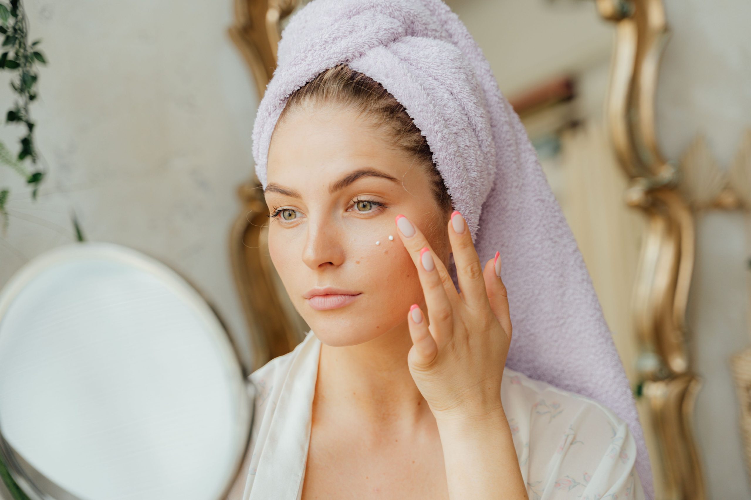 Woman with head towel applying moisturizer in front of mirror indoors. Femme avec une serviette sur la tête appliquant une crème hydratante devant un miroir à l'intérieur.