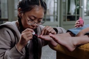 Young girl applying pink nail polish indoors, close-up on pedicure with eyeglasses. Jeune fille appliquant du vernis à ongles rose à l'intérieur, gros plan sur une pédicure avec des lunettes.
