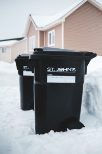 Identical black plastic garbage cans with white inscriptions near snowdrift and residential house exterior in suburb in winter under serene sky Des poubelles en plastique noir identiques avec des inscriptions blanches près d'une congère et de l'extérieur d'une maison résidentielle en banlieue en hiver sous un ciel serein