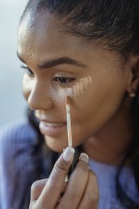 Crop content African American female applying concealer on lower eyelid by using applicator and looking away Contenu recadré d'une femme afro-américaine appliquant du correcteur sur la paupière inférieure à l'aide d'un applicateur et regardant ailleurs